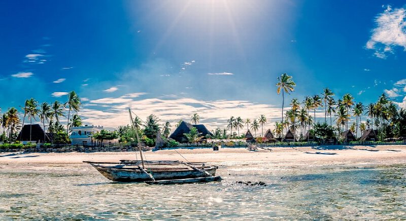 A wooden dhow moored in clear turquoise shallows in front of palms and thatched beach buildings in Zanzibar